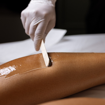 A close-up of a person's leg with an oily skin treatment, while another person wearing a white glove uses a wooden spatula to apply or spread the treatment. The background is a clean white table with a dark, professional setting.