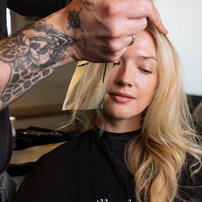 A woman with long blonde hair sits while a tattooed stylist uses a black comb to section her hair. She wears a black salon cape with the text 'milk + honey' visible near the bottom.