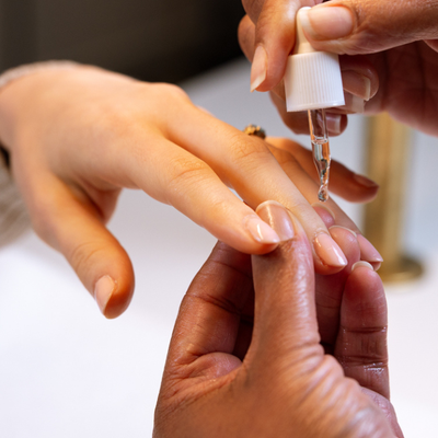 A close-up of a nail care dropper being used to apply oil to a fingernail, showing hands and a white dropper bottle.