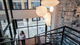 A modern interior staircase with black metal railings, wooden steps, large white ribbed pendant lights, and a patterned wall on the right. A woman is walking down the stairs near the left side.