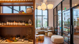 A modern wellness or spa retail interior with wooden shelving displaying many small bottles and jars, a curved seating area with cushions near large windows, pendant lights, and round ottomans on a wood floor.