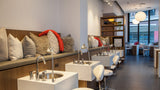 A modern spa or salon interior with multiple white sink stations, chrome faucets, white stools, a wood-and-cushion seating area with pillows, and large windows in the background.