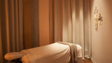 A massage table covered in white linens with a beige throw blanket, set in a warmly lit spa room with draped curtains and wood flooring.