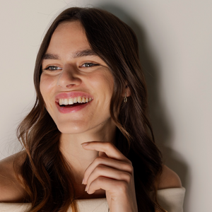 A smiling young woman with long wavy brown hair leans against a plain light beige wall, wearing a small gold hoop earring and a cream off-the-shoulder top.