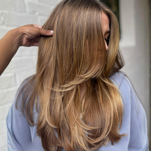 Long, layered wavy hair in a warm blonde balayage color, shown on a model from the back with a hand lifting a section of the hair to show texture and shine.