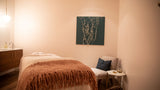A spa-style treatment room featuring a massage table with a rust-colored fringed throw blanket, a white lounge chair with a blue accent pillow, and a dark teal abstract wall art piece hanging on the beige wall.