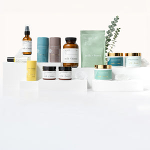 A collection of skincare and beauty products from the brand 'naturally beautiful' arranged on white display pedestals against a clean white background. The products include various bottles, jars, and boxes in muted teal, brown, yellow, and white, with a small green eucalyptus sprig as a decorative prop.