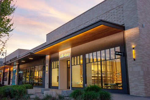 Modern commercial storefront building with large glass windows, warm interior lighting, a wood-look overhang, and a light stone exterior at dusk.