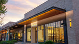 Modern commercial storefront building with large glass windows, warm interior lighting, a wood-look overhang, and a light stone exterior at dusk.