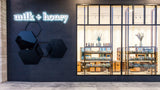 Milk + Honey storefront exterior with a dark gray wall, glowing white neon-style logo, black hexagonal wall decor, and large glass windows showing shelves inside.