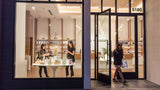 A storefront window display with large black-framed glass doors and windows, showing a warmly lit interior shop. Two women stand inside near a small display table, and one woman walks out through the open door.