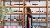 A woman walks past tall wooden shelves stocked with small bottles, jars, and boxes in a bright retail interior.