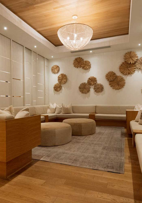 A modern, warm-toned lounge or spa interior with wooden seating, cream cushions, a round glass chandelier, wood ceiling panels, and decorative woven wall pieces.