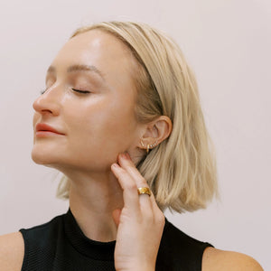 Woman wearing gold earrings and a ring, touching her neck against a plain background