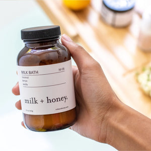 Hand holding a bottle of 'Milk + Honey' milk bath with a blurred background