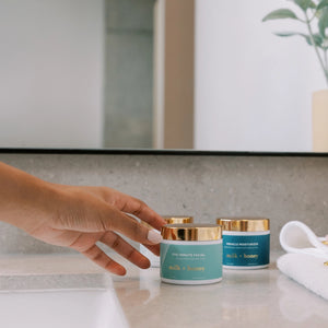 Hand reaching for a skincare product on a bathroom counter with a mirror and towel in the background.