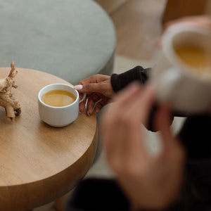 Two people sitting at a table with coffee cups, one of which is being held.