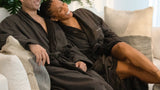 Two people in dark robes sitting on a couch with plants in the background