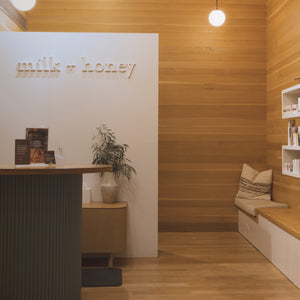 A modern interior space with warm wood paneling and flooring, a white reception desk, floating wall shelves with products, and a bench with cushions. The wall displays the text 'milk + honey'.