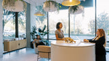 A modern interior space with large windows, hanging pendant lights, and several people gathered around a round white reception desk. The scene includes a woman seated on a chair near the window, two standing women at the desk, and decorative plants throughout the room.