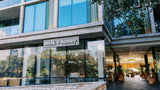 Modern glass-front building exterior with a visible 'milk + honey' sign above the entrance, reflections of trees in the windows, and a paved walkway leading into the lobby.