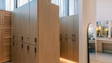 A modern interior space with tall light wood storage lockers and cabinets, a full-length floor mirror, and a vanity area in the background.