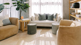 A styled living room scene featuring a cream sofa with sage green and white pillows, a tan armchair, a matching tan loveseat, two gray cylindrical ottomans, a wooden side table with books and a small plant, and a wooden shelving unit in the background.