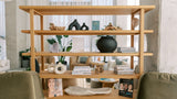 A light natural wood open shelving unit styled with home decor, books, and plants in a modern living room setting.