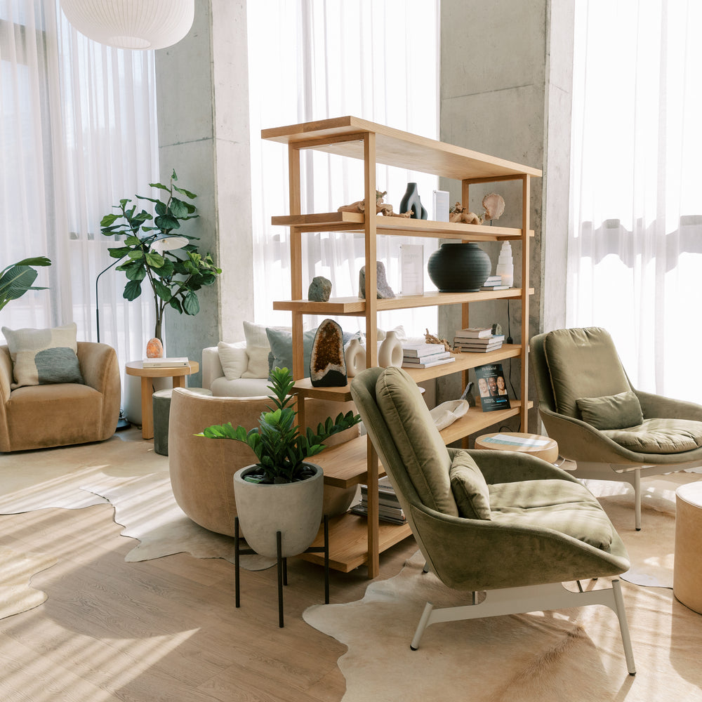 A modern, sunlit living room featuring a tall wooden bookshelf styled with home decor, surrounded by two green upholstered armchairs, beige armchairs, a round beige side table, and large potted plants.
