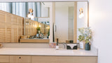 A modern bathroom vanity with a light wood console table, a white countertop, a large wall mirror, and warm wall sconces. A vase with white flowers sits on the right side of the counter.