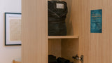 A light wood wardrobe or cabinet with an open door, showing a dark gray bag on the top shelf, black slide sandals on the bottom shelf, and a small blue 'reward' card on the door.