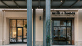 Front exterior of a storefront with the sign 'milk + honey spa | retreat | shop' above glass doors, framed by large gray columns and a covered walkway with hanging lights.