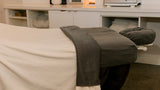 A massage table in a spa setting, covered with a white sheet and a dark gray towel, with a matching headrest. In the background, a white treatment counter holds a digital clock, a lit candle, and spa equipment.