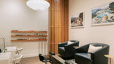 A modern spa or salon interior with two black leather reclining chairs, a pedicure treatment table, white vanity tables, wall shelves, framed artwork, and a slatted wood screen.