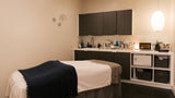 A spa treatment room featuring a massage table with white and navy blue linens, a built-in treatment counter with shelves and cabinets, and a hanging pendant light.