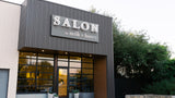 A modern storefront with a dark gray corrugated metal facade and a large sign reading 'SALON by milk + honey' above glass entrance doors, with trees and a pale sky in the background.