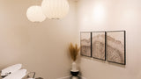 A modern interior corner with two white spherical pendant lights hanging from the ceiling, cream walls, three framed abstract wood-art prints, a black-and-white pedestal, and a vase with dried grass. No people or animals are present.