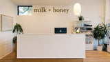 A bright, modern reception desk in a wellness or skincare shop with the words 'milk + honey' displayed on the wall above it. The white desk has a black laptop on top, with shelves, potted plants, and a hanging pendant light in the background.