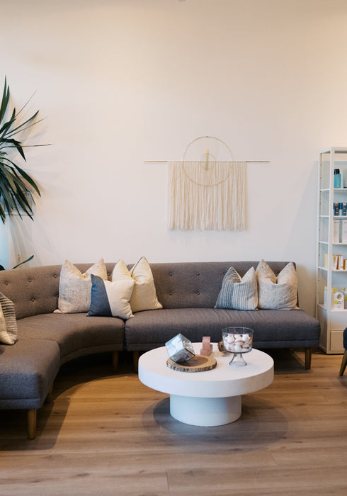 A modern living room setup featuring a curved gray fabric sofa with beige and blue-gray pillows, a round white coffee table, two dark blue armchairs with yellow pillows, a white shelving unit filled with products, and a large window with black frames.