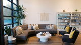 A modern living room setup featuring a curved gray fabric sofa with beige and blue-gray pillows, a round white coffee table, two dark blue armchairs with yellow pillows, a white shelving unit filled with products, and a large window with black frames.