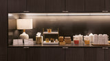A modern beverage and snack bar counter set against dark wood cabinetry, featuring a white ceramic table lamp, stacks of paper cups, small jars, and boxes under warm under-cabinet lighting.