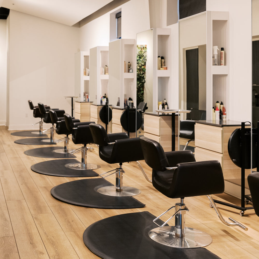 A salon interior with multiple black swivel styling chairs arranged on a light wood floor in front of mirrors and built-in shelving.