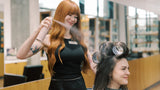 A woman with long auburn hair stands in a salon while another woman sits in the chair with curlers in her hair, receiving a hair spray treatment.