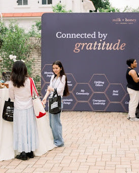 Three women standing outdoors in front of a large dark blue banner that reads 'Connected by gratitude' with honey-themed text and geometric patterns. One woman holds a white tote bag with red accents, and another carries a black shoulder bag.
