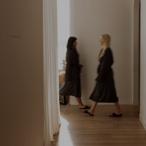 A dimly lit interior hallway with two women walking in the distance. The scene has a dark overlay and a small padlock icon near the upper left.