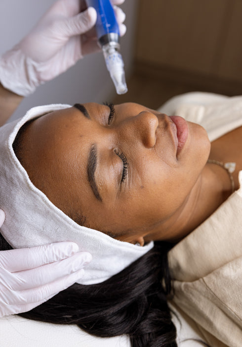 A close-up lifestyle shot of a woman receiving a facial treatment, with a gloved hand holding a blue syringe above her forehead while another hand adjusts a white towel on her head.