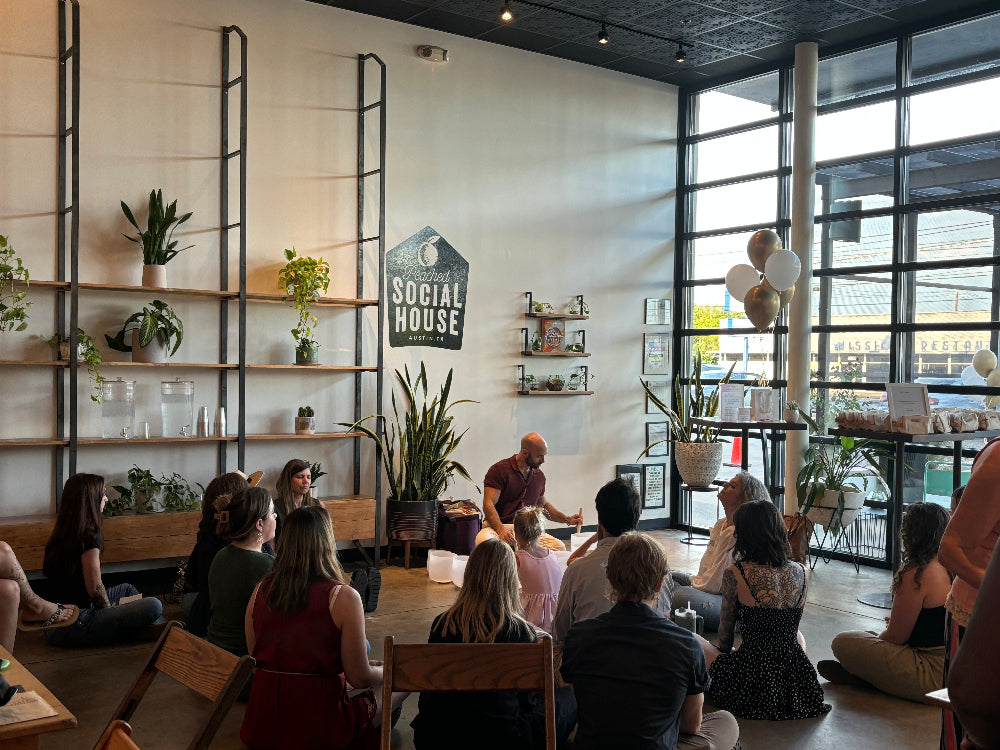 A group of people sitting on the floor in a bright, modern cafe or workshop space, facing a presenter who is standing near a white wall with shelves and a sign that reads 'Social House'.