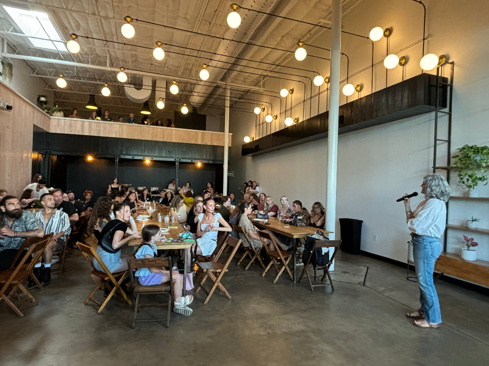 A group of people seated at tables in a bright loft-style restaurant or event space, with a woman standing on the right holding a microphone in front of an open loft ceiling with hanging globe lights.