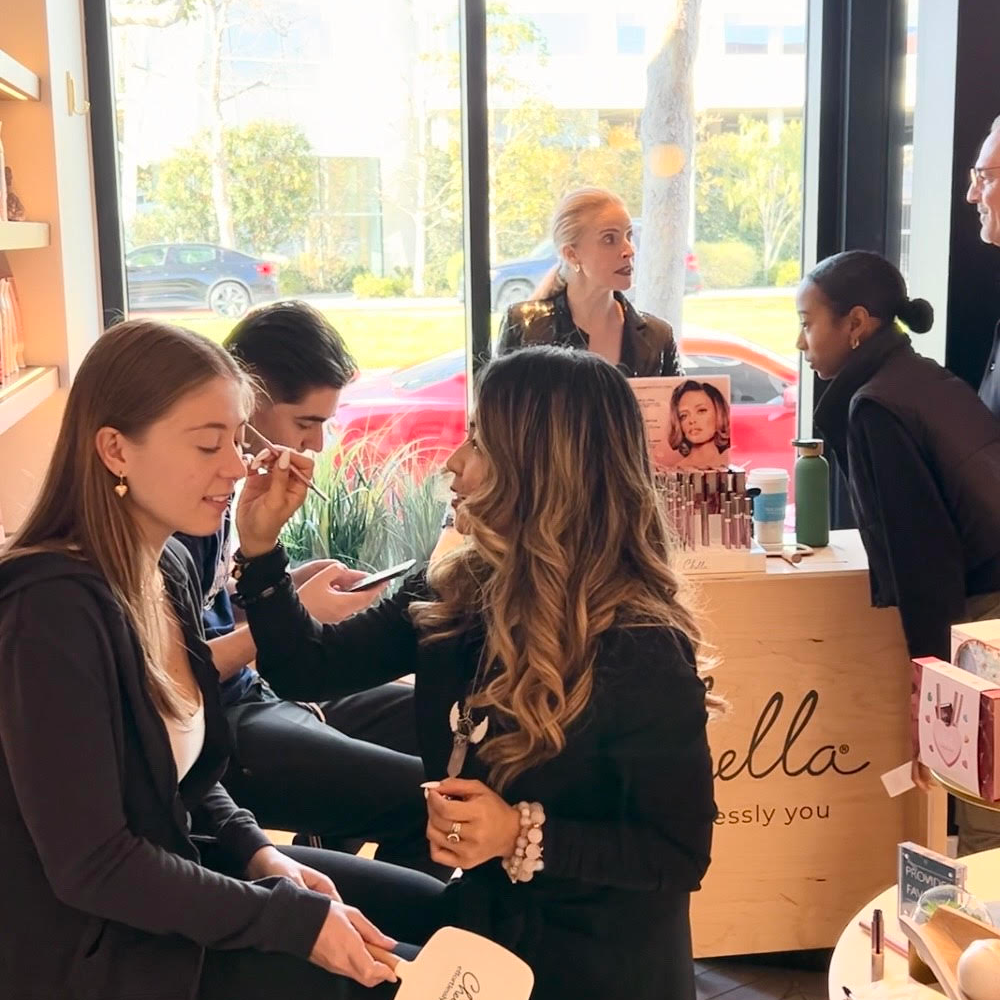 A woman in a black coat and black boots stands in a beauty salon or makeup studio, holding a small white card while another woman applies makeup to her face. Other people and a counter with the brand name 'ella' are visible in the background near large windows.