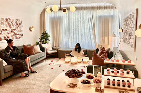 A cozy, upscale lounge or gift lounge setting with two women sitting on sofas, a coffee table with bowls of dried fruits and candles, and a display shelf of small pink and red packaged items. The room features warm neutral tones, beige curtains, and modern wall art.